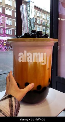 Die Hand hält eine riesige Teetasse mit schwarzen Tapioka-Perlen und klarem Stroh in der Nähe des Fensters Stockfoto