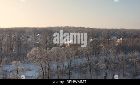 Luftaufnahmen von Drohnen fliegen über ein kleines Dorf mit Häusern und bewegen sich in Richtung eines riesigen, dichten Waldes. Bäume und Boden sind mit frischem weißem Schnee bedeckt Stockfoto