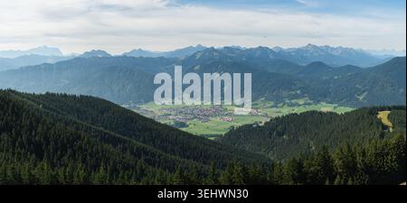 Ein atemberaubender Panoramablick zeigt die tiefen, bewaldeten Täler und sanften Hügel, die zu den majestätischen Gipfeln der Zugspitze und des Voral führen Stockfoto
