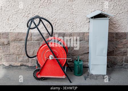Roter Feuerlöschschlauch mit Gießkanne in der Nähe einer strukturierten Wohnwand im städtischen Außenbereich Stockfoto