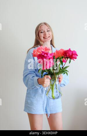 Glückliche blonde Frau mit bunten Pfingstrosenstrauß in hellem Studio, femininer Schönheit und Selbstpflegekonzept mit natürlichem Licht. Stockfoto
