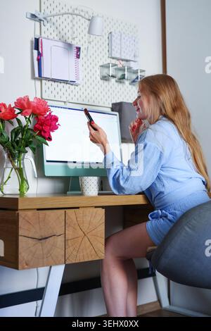 Entspannte junge Frau mit Kaffeetasse im stilvollen Home Office, lässige Indoor-Lifestyle-Szene mit Computer und frischen Pfingstrosen. Stockfoto