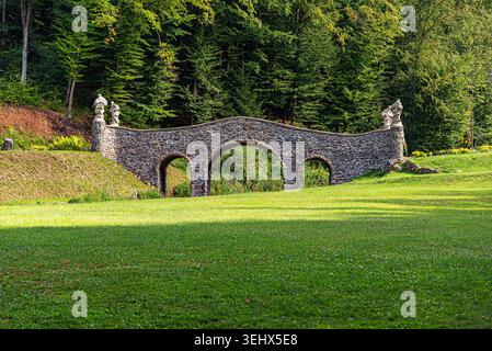 Steinbrücke mit drei Bögen steht auf einem üppig grünen Rasen vor einem dichten Wald. Stockfoto