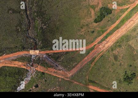 Aus der Vogelperspektive auf roten, unbefestigten Straßen, die sich durch grüne Savannenlandschaft mit einem kleinen Bach in Waterberg District Municipality, Limpopo, Südafrika, schlängeln. Stockfoto