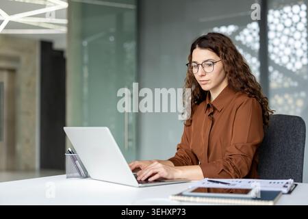 Junge Geschäftsfrau mit lockigem Haar und Brille, die an einem Schreibtisch sitzt, auf einem Laptop schreibt und sich auf ihre digitale Arbeit in einer modernen Büroumgebung konzentriert Stockfoto
