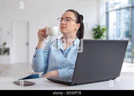Geschäftsfrau genießt eine Kaffeepause neben einem Laptop in einem hellen Büroarbeitsplatz, der für ruhige Remote-Arbeit und modernen professionellen Lebensstil steht Stockfoto