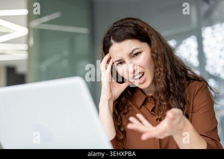 Frustrierte Frau mit lockigem Haar, die Wut und Stress ausdrückt, während sie mit einem technischen Problem auf ihrem Laptop zu kämpfen hat und Kopfschmerzen während einer schwierigen Büroaufgabe hat Stockfoto