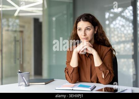 Geschäftsfrau blickt weg, legt ihre Hände in einem modernen Büro auf einen Schreibtisch, tief in Gedanken, konzentriert sich auf geschäftliche Herausforderungen und denkt über Lösungen nach Stockfoto