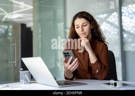 Frau, die sich auf Inhalte auf ihrem Smartphone konzentriert, an einem weißen Schreibtisch mit Laptop- und Stifthalter sitzt und geschäftliche Kommunikation und Mobiltechnologie in einer Unternehmensumgebung repräsentiert Stockfoto