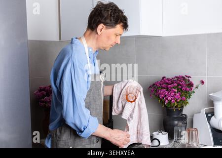 Ein Mann steht an einem Waschbecken in einer Küche und wäscht schmutziges Geschirr und Gläser. Blumen sitzen auf der Theke, während frisches Licht durch das Fenster hereinkommt. Die se Stockfoto