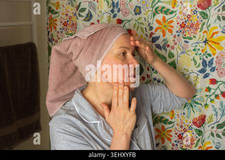 Eine 50-jährige Frau bereitet ihre Haut vor, indem sie Creme im Badezimmer aufträgt. Sie trägt ein Handtuch auf dem Kopf und steht vor einem bunten Duschcurtai Stockfoto