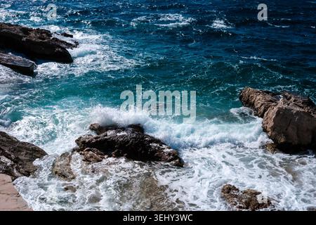 Mächtige Meereswellen krachen mit weißem Schaum gegen dunkle, verwitterte Felsen entlang einer zerklüfteten Küste bei hellem Tageslicht. Stockfoto