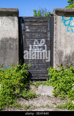 Graffiti-Künstler haben dieses verwitterte Holztor in einer Betonwand mit Tags wie „BOTOK“ und „UHU33“ geschmückt. Ihre urbanen Ausdrucksformen stehen im Kontrast Stockfoto
