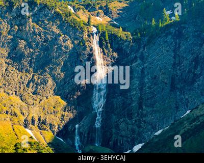 Ein hoher Wasserfall stürzt über eine zerklüftete felsige Klippe in ein grünes alpines Tal, eingerahmt von Kiefern und Schneeflächen, mit warmen Sonnenlichtangaben Stockfoto