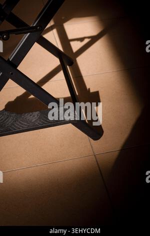 Metallgestell und gemusterte Fußstütze eines Klappstuhls schaffen starke geometrische Formen und Schatten auf hellbraunen Bodenfliesen Stockfoto