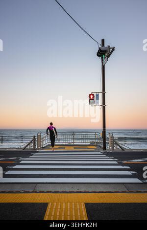 Blick auf einen Surfer, der ein Surfbrett über eine Fußgängerüberquerung in Richtung Meer trägt, unter einem weichen Sonnenuntergangshimmel Kamakura, Kanagawa, Japan. Stockfoto
