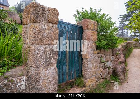 Ein altes, verwittertes blaues Holztor befindet sich zwischen Säulen und einer Trockenmauer aus rosa Granitblöcken. Ein Schotterweg schlängelt sich an ihm, flankiert von Stockfoto