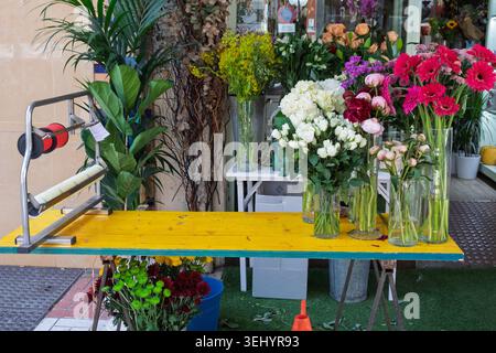Frisch geschnittene Blumen sind in Vasen auf einem gelben Tisch an einem Blumenstand am Straßenrand angeordnet Stockfoto