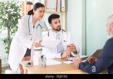 Senior man Kunde der medizinischen Klinik sitzt gegenüber männlichen und weiblichen Arzt und kommuniziert mit Spezialisten Stockfoto
