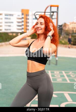 Lächelnde rothaarige Athletin, die vor dem Workout kabellose Kopfhörer aufsetzt. Glückliche Sportlerin, die im Fitnessstudio im Freien die Kamera ansieht Stockfoto