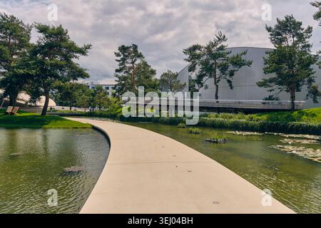 Wolfsburg, Deutschland - 06. Juni 2025: Blick auf den Autostädter Park mit einem geschwungenen Gang über einen Teich mit Seerosen, grünen Bäumen und modernem Archit Stockfoto