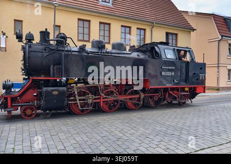 Bad Doberan, Deutschland - 21. Juli 2025: Blick auf die Dampflokomotive 99 2322-8 der Molli Bahn auf den Straßengleisen mit Fahrer im Fahrerhaus und historischer Bu Stockfoto