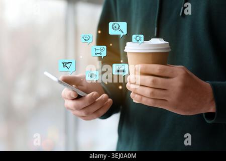 Digitale Symbole und ein Mann mit einer Tasse Kaffee und ein Mobiltelefon in der Nähe des Fensters. Konzept des digitalen Marketings Stockfoto