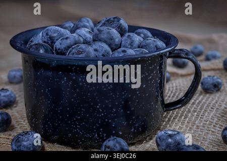 Eine seitliche Nahaufnahme eines dunklen gesprenkelten Emailbechers, der bis zum Rand mit frischen Bio-Heidelbeeren gefüllt ist und auf einem rustikalen Sacklappen mit verstreuten Stockfoto