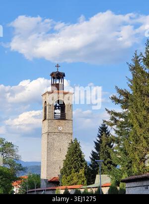 Ein historischer Kirchturm aus Stein mit einer Uhr und einem hölzernen Glockenturm erhebt sich über Bäumen vor einem klaren blauen Himmel und hebt das traditionelle Mauerwerk hervor Stockfoto