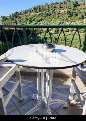 Sonnendurchfluteter Marmortisch auf einem Balkon mit silbernem Aschenbecher und zwei weißen Stühlen mit Blick auf die üppig grünen Hügel. Ruft Entspannung, Morgenkaffee, Quie hervor Stockfoto