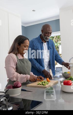 Diverse Paare, die Zucchini auf der Kücheninsel hacken, Frau in olivgrüner Schürze, Mann, der Produkte spült Stockfoto