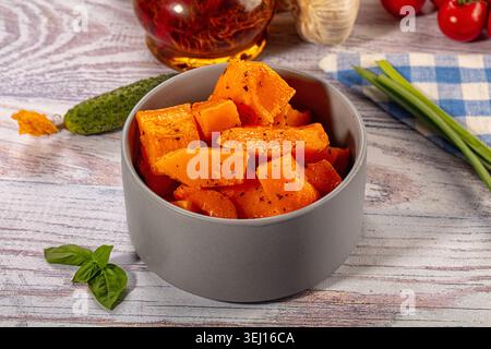 Gebackene, orangene Kürbisstücke, gewürzt mit Gewürzen in einer grauen Keramikschale. Gesundes vegetarisches Gericht auf hellem Holzhintergrund mit frischem Gemüse wie Bas Stockfoto