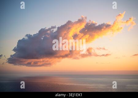 Große dramatische Cumulus-Wolke, beleuchtet in lebendigen Orange- und Goldtönen vor sanftem pastellfarbenen Sonnenuntergang über dem friedlichen mediterranen Horizont in der Nähe Stockfoto