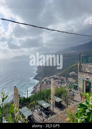 Atemberaubende Aussicht von einer Bergterrasse mit Blick auf ein malerisches Dorf am Meer. Leere Tische mit Sonnenschirmen bieten eine ruhige Atmosphäre. Wolken und Stockfoto