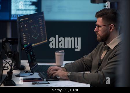 Kaukasischer junger erwachsener Mann, der Cybersicherheitsdaten auf mehreren Bildschirmen analysiert und im Büro auf einem Laptop schreibt. Seitenansicht, ideal für IT-Sicherheit, Datenüberwachung und Risikomanagement-Marketing Stockfoto