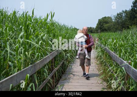 Ein älterer Mann hält ein Kind in den Armen und läuft auf einem Naturlehrpfad. Stockfoto