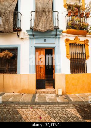 Blick auf eine traditionelle andalusische Hausfassade mit offener Holztür, dekorativen blauen und gelben Fensterrahmen und Blumentöpfen auf Balkonen in Cord Stockfoto