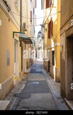 Piran, Slowenien. Mai 2026. Eine enge, sonnendurchflutete Gasse, gesäumt von hohen, gelben Gebäuden. Die Straße ist mit Stein und Asphalt und Uti gepflastert Stockfoto