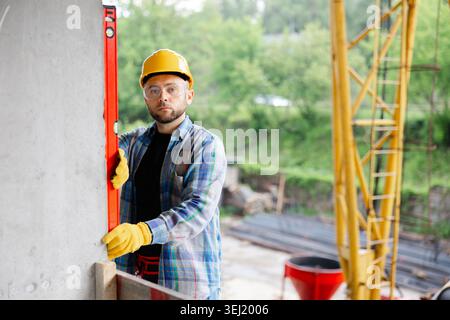 Fachkundiger Bauarbeiter, der Sicherheitsausrüstung trägt und die vertikale Ausrichtung einer Wand auf einer Baustelle mit einer Wasserwaage überprüft Stockfoto