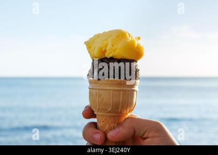 Nahaufnahme einer Hand, die eine Eiskugel mit Mango- und Schokoladenlöffeln vor einem ruhigen Meereshorizont hält. Stockfoto