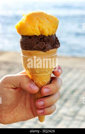 Nahaufnahme einer Hand, die eine Eiskugel mit Schokoladen- und gelben Sorbetlöffeln in der Nähe des Meeres hält. Weichzeichner Stockfoto