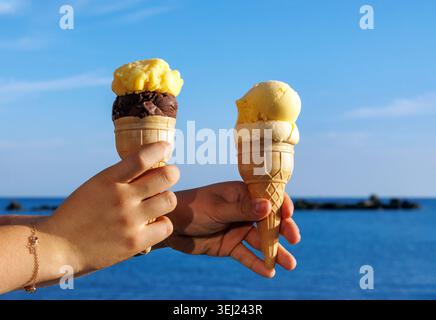 Nahaufnahme von zwei Händen, die Eiskegel vor blauem Meer und Himmel halten. Stockfoto