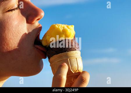 Eine Frau isst eine Eiskugel mit Schokolade und gelbem Mangobrüchsorbet vor einem hellblauen Himmel. Stockfoto