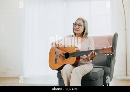 Gesunde ältere asiatische Musikerin spielt Gitarrenmusik Liebhaber im Wohnzimmer drinnen zu Hause. Stockfoto