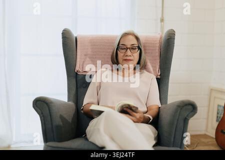 Seniorin, die Buch liest, Sitzen, entspannen Sie sich ruhig zu Hause in sanftem Sonnenlicht. Asiatische ältere Brille Happy Smile. Bequemer Stuhl hält das Buch entspannt p Stockfoto