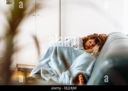 Die junge Frau liegt auf einem Ledersofa, das mit einer warmen, gemütlichen Decke bedeckt ist Stockfoto