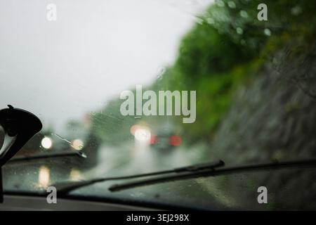Blick von der Innenseite des Armaturenbretts des Fahrzeugs, Blick durch eine regnerische, nasse Frontscheibe mit Wischerblättern und verschwommenen Scheinwerfern des Straßenverkehrs während des Tages Stockfoto
