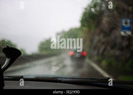 Ein Innenraum-POV-Bild eines Armaturenbretts und einer Windschutzscheibe aus nassem Glas, die während eines Sturms von Regentropfen bedeckt war, gefolgt von einem anderen Fahrzeug auf einem schmalen Hang Stockfoto
