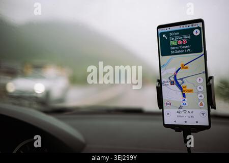 Eine Nahaufnahme eines Mobiltelefons in einem Autohalter, das GPS-Navigation mit Wegbeschreibungen an einem bewölkten Tag anzeigt, mit verschwommener Autobahn und Autohea Stockfoto