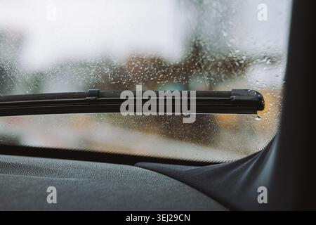Eine Nahaufnahme aus dem Armaturenbrett eines Fahrzeugs mit Blick auf ein schwarzes Wischerblatt, das auf einer regenbedeckten Windschutzscheibe mit einem verschwommenen Straßenrand geparkt ist Stockfoto